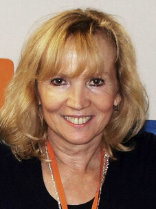 Monika Seifer mit blondem Haar und dunkler Kleidung, lächelnd vor neutralem Hintergrund.